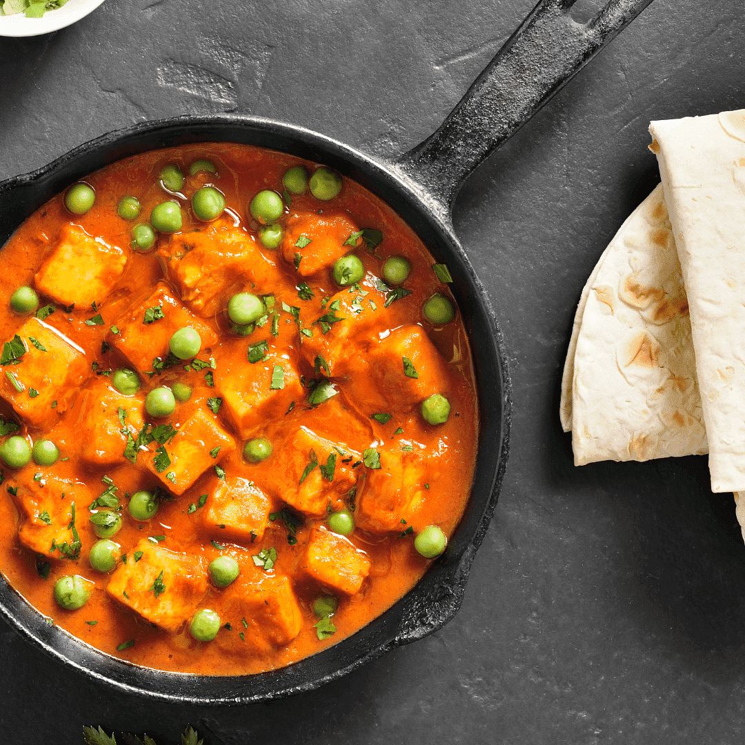 Pan of paneer tikka masala curry with peas next to a stack of naan bread on a dark surface.