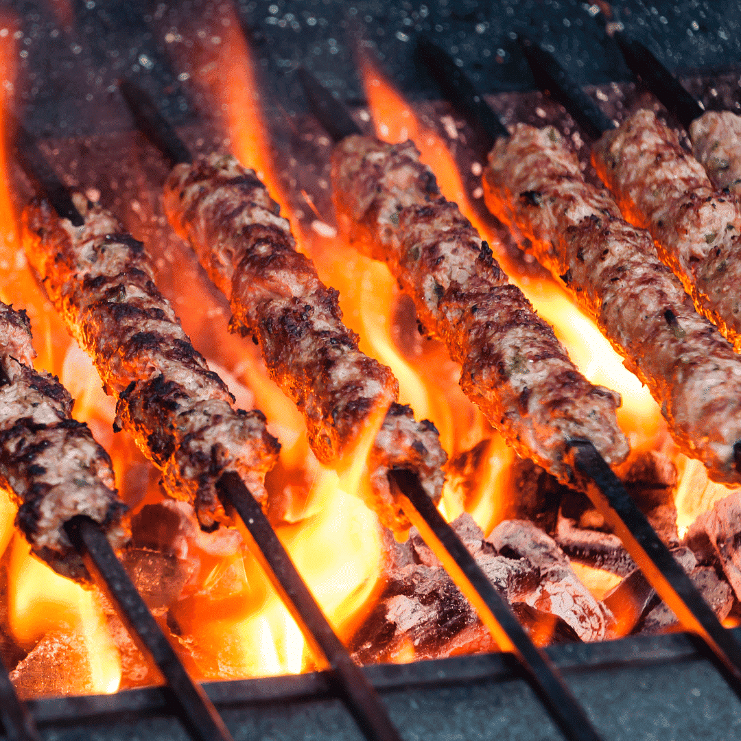 Skewers of meat cooking over a hot charcoal fire, with flames rising between the skewers.