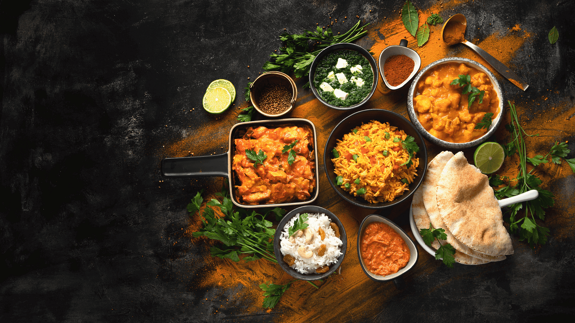 An array of Indian dishes including rice, curry, naan, and various spices on a dark background.