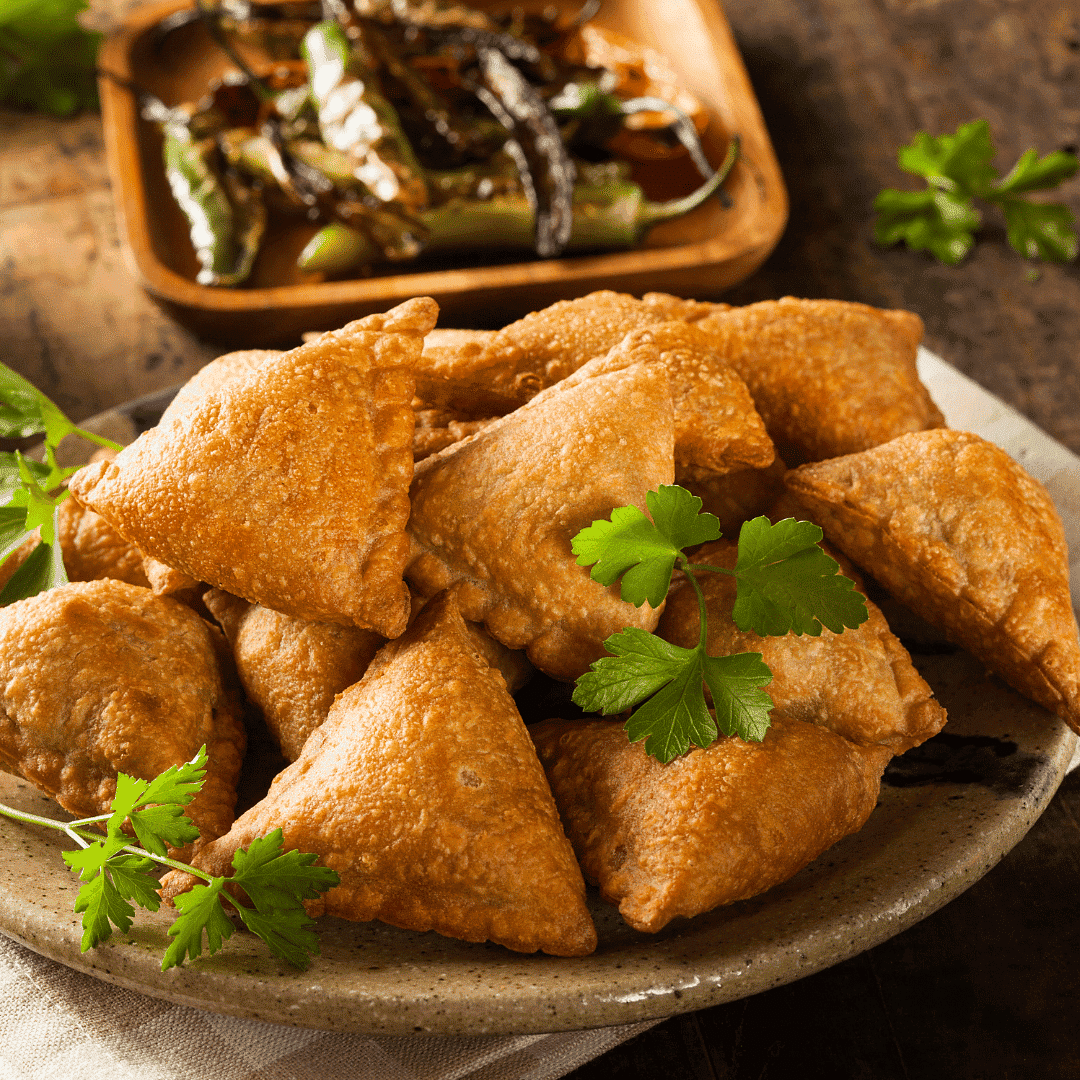 Plate of golden-brown samosas with fresh cilantro garnish, side of fried green chilies in background.